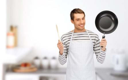 دانلود افراد ، مفهوم آشپزی و آشپزی – مرد خوشحال یا آشپزی در پیش بند با ماهی تابه و قاشق چوبی بر روی زمینه آشپزخانه منزل