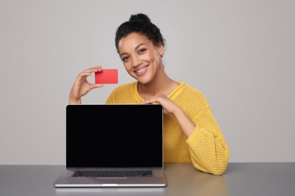دانلود لبخند نژاد مختلط آفریقایی آمریکایی – زن قفقازی که صفحه نمایش رایانه لپ تاپ سیاه و کارت اعتباری خالی را نشان می دهد ، روی میز over نشسته است