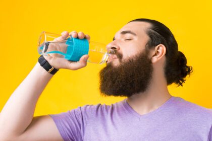 دانلود عکس از نزدیک عکس مرد ریشو در حال نوشیدن آب از شیشه بطری روی پس زمینه زرد