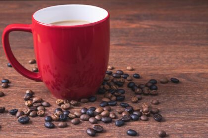 دانلود عکس قهوه در فنجان قهوه قرمز در کنار دانه های قهوه ریخته شده روی میز چوبی