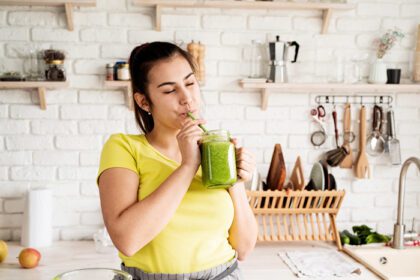 دانلود عکس زن جوان سبزه در حال نوشیدن اسموتی سبز در آشپزخانه