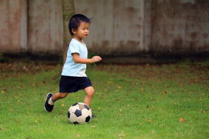 دانلود عکس پسر آسیایی در حال بازی فوتبال در پارک بچه دریبل زدن توپ