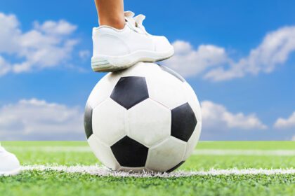 دانلود عکس پاهای پسری با کفش های کتانی سفید که در وسط زمین فوتبال روی توپ فوتبال پا می گذارد