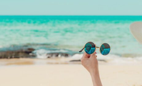 دانلود عکس زنی که عینک آفتابی را در دست گرفته و روی ساحل شنی نشسته است