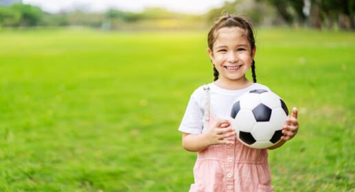 دانلود عکس دختر بچه خندان که توپ فوتبال در دست دارد و در سبزه ایستاده است