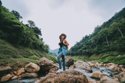دانلود عکس زن آسیایی مسافر با کوله پشتی در حال لذت بردن از منظره رودخانه کوهستان و منظره جنگل در طول تعطیلات